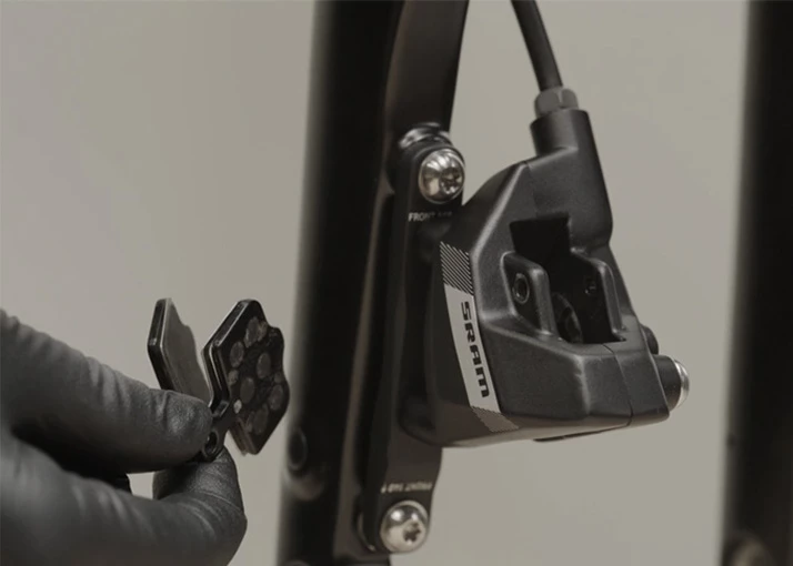 自転車のフレームに取り付けられたSRAM製の自転車用ブレーキキャリパーの横で、ディスクブレーキパッドを手に持つ手袋をはめた手。.
