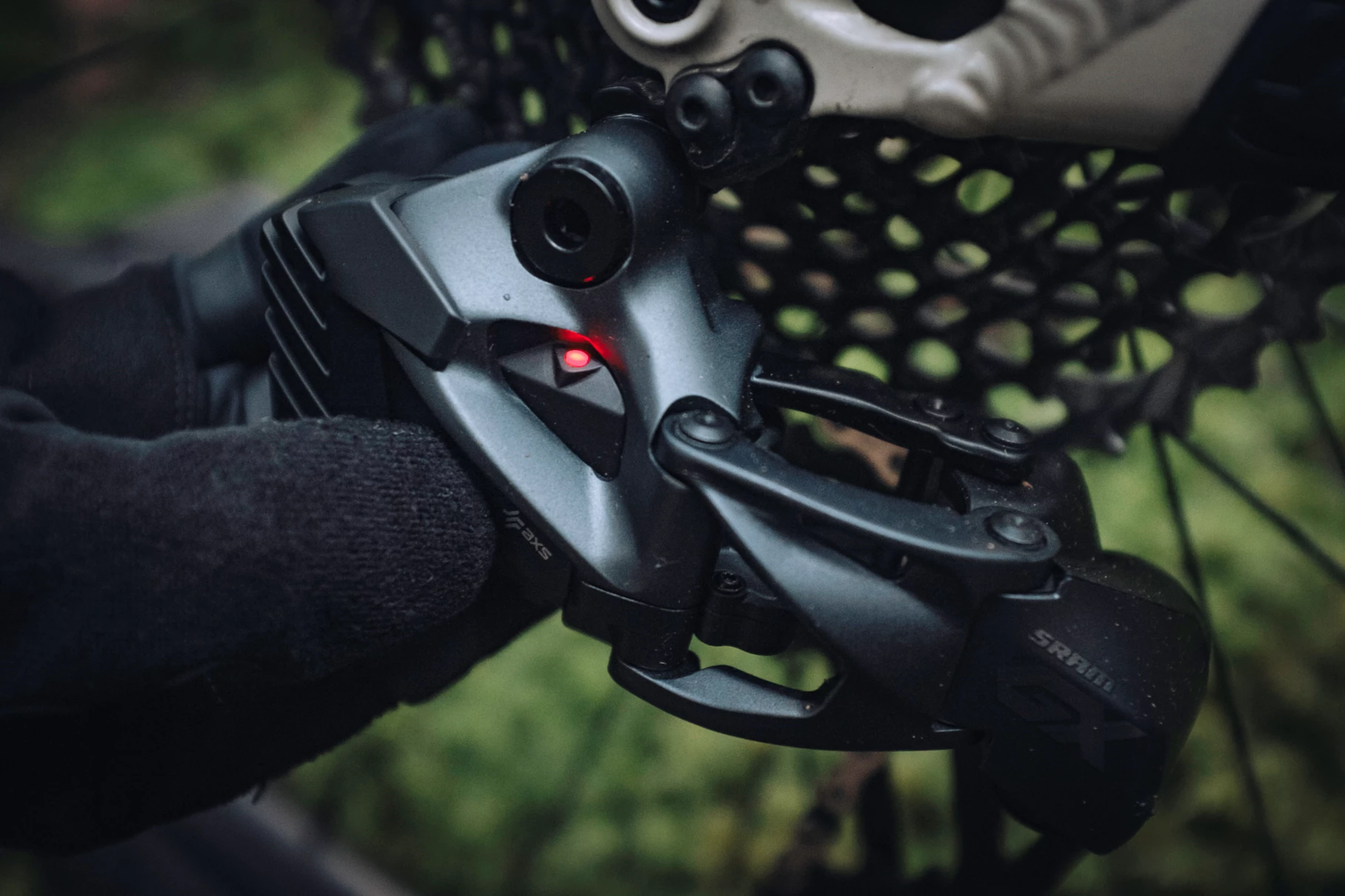 手袋をはめた手が、小さな赤いステータスランプが点灯しているSRAM GX AXSのマウンテンバイク用リアディレイラーを調整している。.