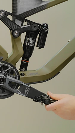 マットなオリーブグリーンのマウンテンバイクのペダルに、手が触れている。この自転車にはリアサスペンションが装備されている。.