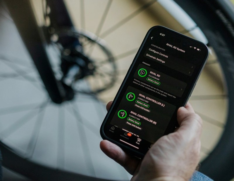 SRAM AXSアプリが表示されたスマートフォンを手に持ち、Rivalドライブトレインシステムのバッテリー残量や設定情報が表示されている。.
