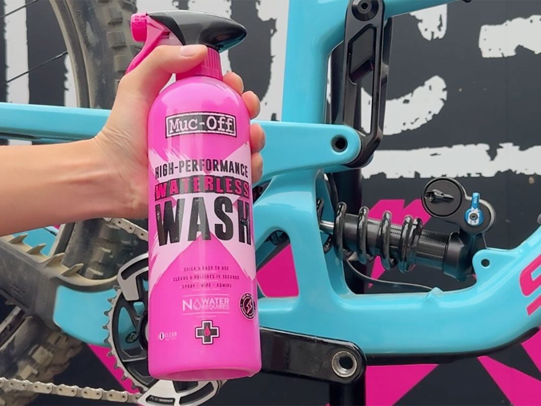 青いマウンテンバイクのフレームを背景に、ピンクのMuc-Off Waterless Washのスプレーボトルを手にした手。.