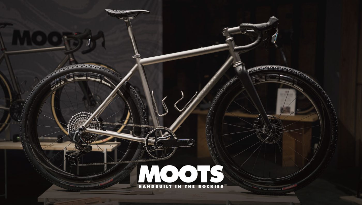 Mootsのロゴの前に置かれた、カーボンフォーク、ノビータイヤ、シングルチェーンリングのドライブトレインを備えたシルバーのMoots製グラベルバイク。.