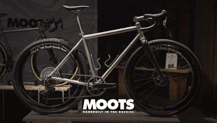 Mootsのロゴの前に置かれた、カーボンフォーク、ノビータイヤ、シングルチェーンリングのドライブトレインを備えたシルバーのMoots製グラベルバイク。.