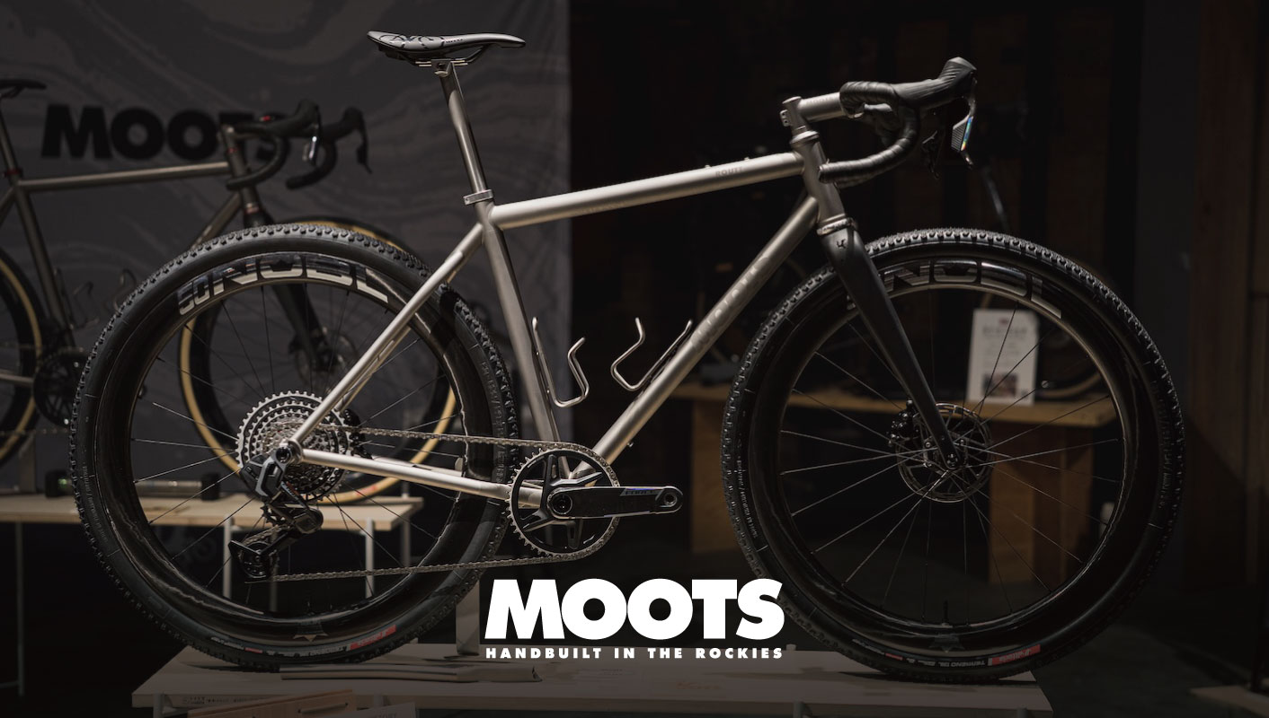 Mootsのロゴの前に置かれた、カーボンフォーク、ノビータイヤ、シングルチェーンリングのドライブトレインを備えたシルバーのMoots製グラベルバイク。.